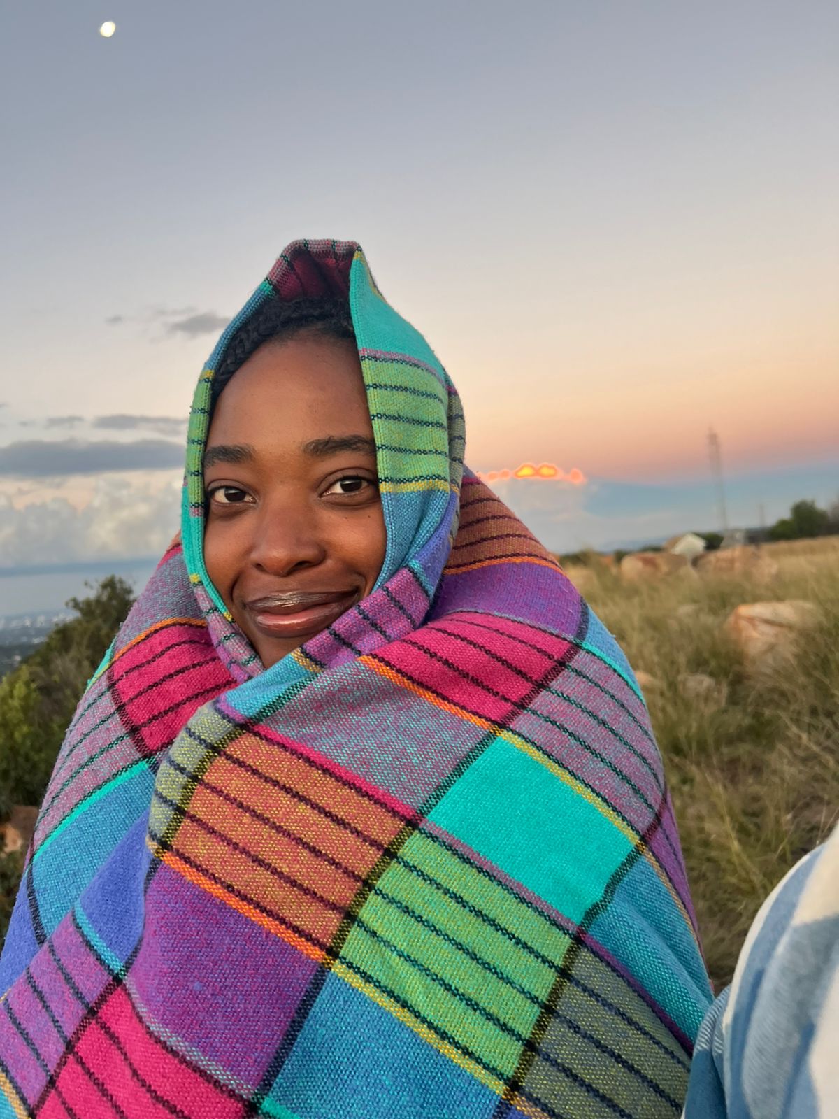 Tshego wrapped in a bright checked blanket on a hillside at dusk, moon overhead
